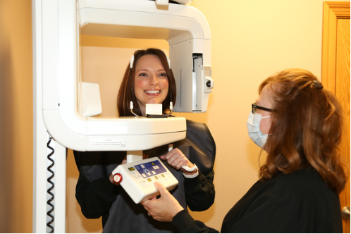 A woman stands in a dental panoramic X-ray machine, while a dental technician in a face mask operates the control panel. The patient is smiling and wearing a protective apron.