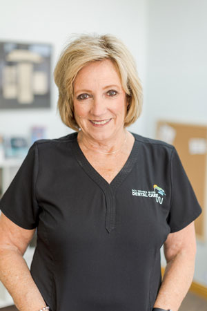 A woman with short blonde hair is smiling and wearing black scrubs with a dental care logo. She is standing in a bright room with a blurry background that includes a bulletin board and shelves with items.