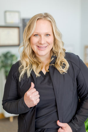 A woman with long blonde hair is smiling at the camera. She is wearing a black jacket and standing in an indoor setting with a plant and some frames visible in the background.