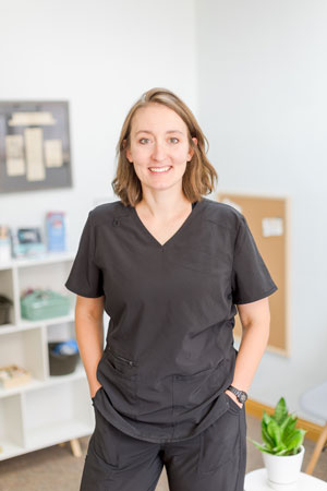 A smiling individual with shoulder-length brown hair stands in a well-lit room, dressed in black scrubs with hands in pockets. Behind them, there is a white bookshelf with various items and pamphlets, a corkboard on the wall, and a small potted plant in the foreground.