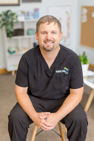 A person with short hair and a beard is sitting on a chair, smiling. He is wearing black scrubs with a logo on the chest. The background shows a bright, organized room with shelves, a bulletin board, and potted plants.