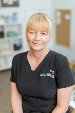 A woman with blonde hair, wearing a black dental care uniform with a logo, is smiling slightly. She is seated in what appears to be a dental office, with shelves and a bulletin board visible in the background.