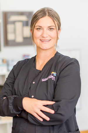 A woman with light brown hair tied back is smiling at the camera. She is wearing a black dental uniform with embroidered text that reads "Hermosa Creek Dental Care" and a small tooth logo. Her arms are crossed in front of her. The background is a dental office.