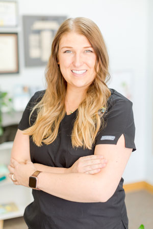 A woman with long, light brown hair is smiling and standing with her arms crossed. She is wearing a black short-sleeved top and a smartwatch. The background includes some framed certificates and a few shelves with items.