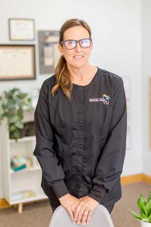 A smiling woman wearing black scrubs and glasses stands in an office. The background shows a bookshelf with books and papers, certificates or framed items on the wall, and a small plant.