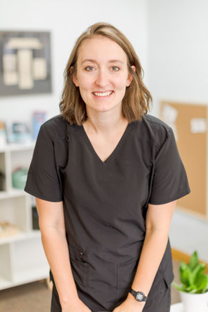 A person with shoulder-length brown hair is smiling at the camera, wearing a black V-neck top. In the background, there are shelves with books and items, and a corkboard on the wall. The setting appears to be an office or workspace.