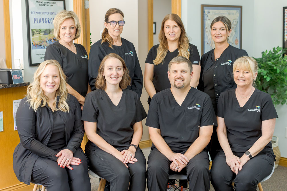 A group of eight people wearing black uniforms pose together in a well-lit room in Des Moines River Dental Care's Carlisle office. Some are sitting while others stand behind them, with a plant and framed pictures adding warmth to the background.