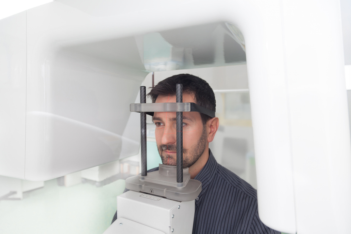 A person is undergoing a panoramic digital X-ray, standing with their chin on a support and head secured by a brace inside a white imaging machine in a clinical setting.