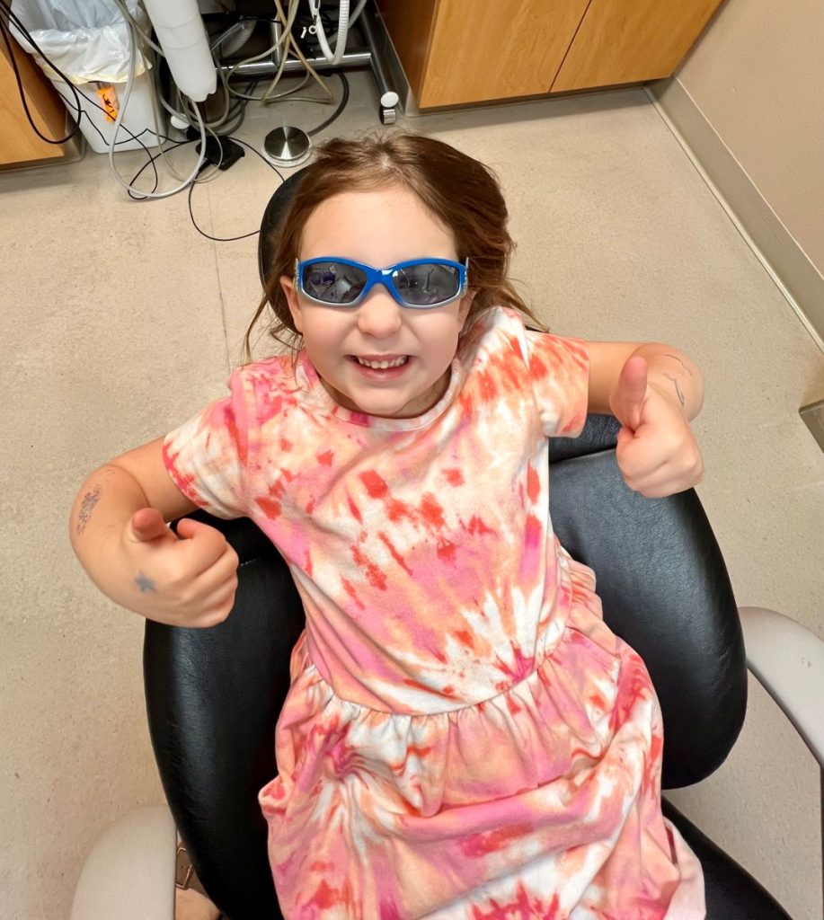 A young girl in a tie-dye dress and blue sunglasses sits in a chair, smiling and giving two thumbs up. The room has beige walls and a cabinet. Cords and a bottle are visible in the background.