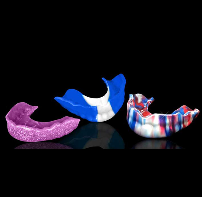 Three colorful custom mouthguards are displayed against a black background. The left one is purple with speckles, the middle is blue and white, and the right one has a red, white, and blue marble pattern.