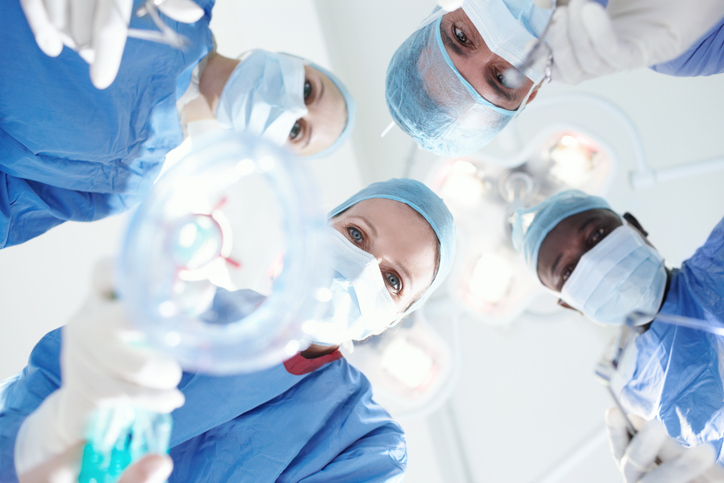 Four surgeons in scrubs and masks look down at a patient on an operating table, with bright surgical lights overhead while one surgeon carefully holds an advanced local anesthesia mask toward the camera.