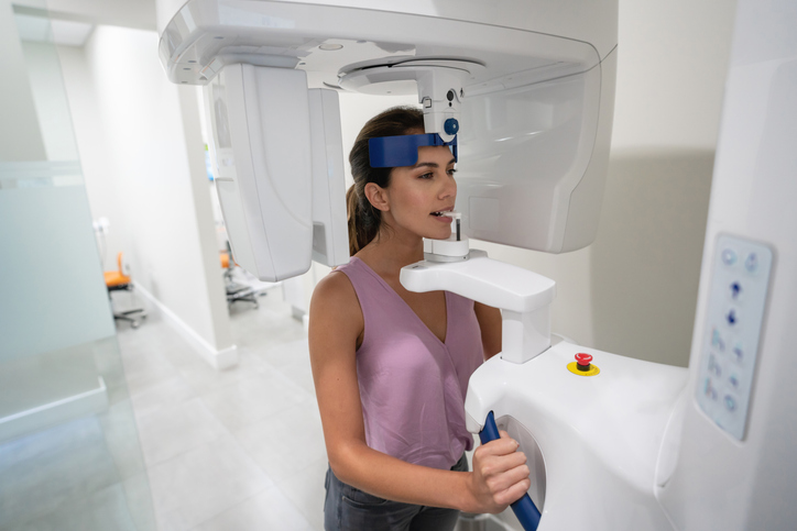 A woman stands in a dental clinic using a panoramic X-ray machine for a dental scan, holding handles and biting a mouthpiece, with her head secured by a blue strap—similar to how CBCT scans are performed for detailed imaging.