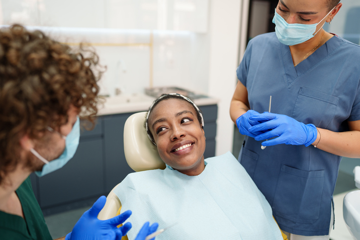 A smiling patient sits in a dental chair after a comprehensive exam, looking at a masked dental professional. Another staff member stands nearby, both wearing gloves and scrubs in a bright, modern clinic setting.