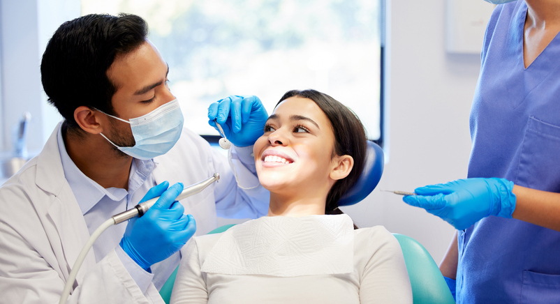 A dentist specializing in comprehensive general dentistry, wearing a face mask, examines a smiling female patient’s teeth as a dental assistant in scrubs and gloves stands nearby holding dental tools in a brightly lit clinic.