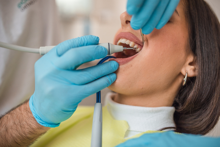 A dentist wearing blue gloves uses dental tools to perform a deep cleaning and examine a patient’s teeth. The patient, in a yellow bib, has her mouth open during the procedure.