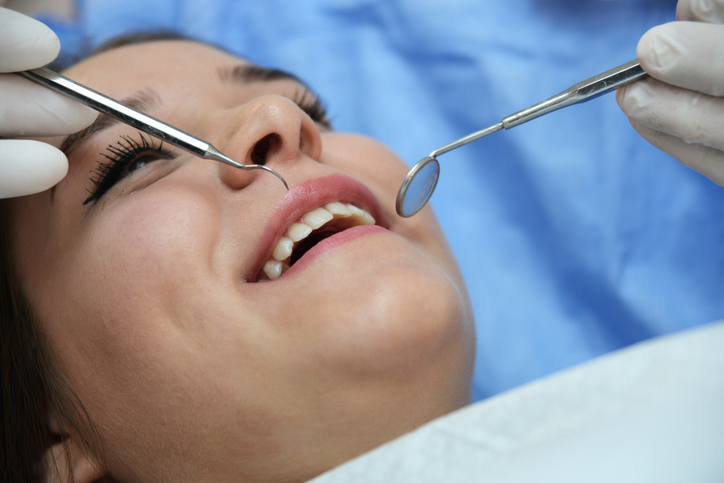 A close-up of a person at a dental checkup, smiling with their mouth open while a dentist examines their teeth using a dental mirror and probe during a routine dental cleaning. The dentist is wearing gloves.