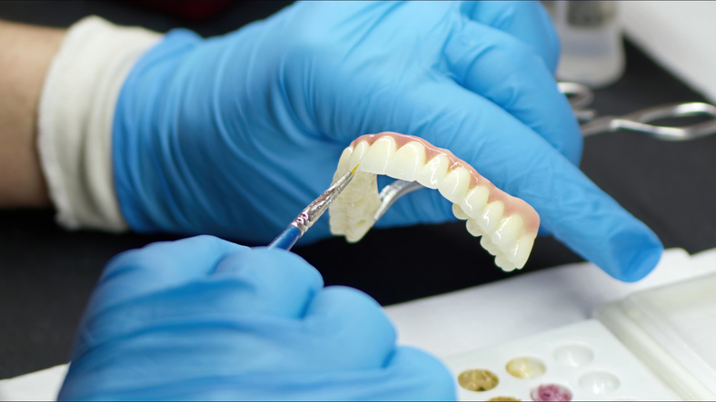 A person wearing blue gloves uses a tool to apply material to dentures or a dental prosthesis, likely in a dental laboratory setting.