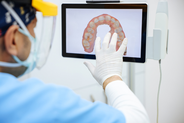 A dentist wearing gloves and a face shield examines digital impressions of a dental arch displayed on a touchscreen monitor.