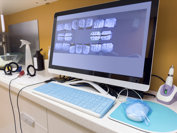 A computer monitor on a dental office counter displays dental X-rays and electronic records. A blue keyboard, mouse, headphones, dental tools, and cleaning supplies are also visible on the countertop.