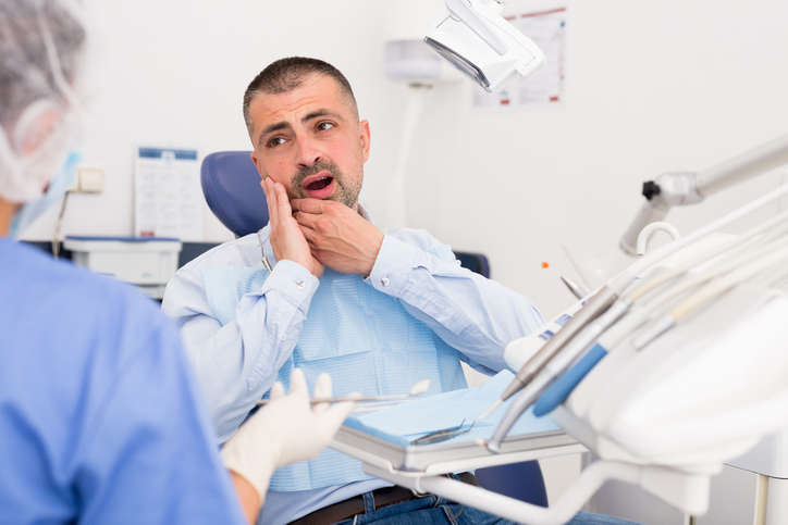 A man sits in a dental chair, holding his cheek in pain and talking to a dentist in scrubs, suggesting he may need emergency dentistry. Dental tools and equipment are visible in the foreground.