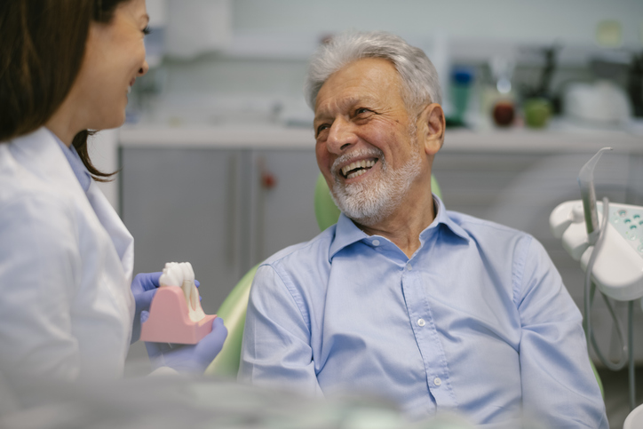 An older man sits in a dental chair, smiling at a dentist who is holding a dental model. The dentist, practicing geriatric dentistry, wears gloves and a white coat in a bright, modern dental office.