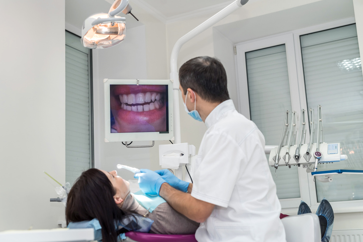 A dentist examines a patient's teeth using an intraoral camera, while an image of the patient's mouth is displayed on a monitor in a modern dental clinic.