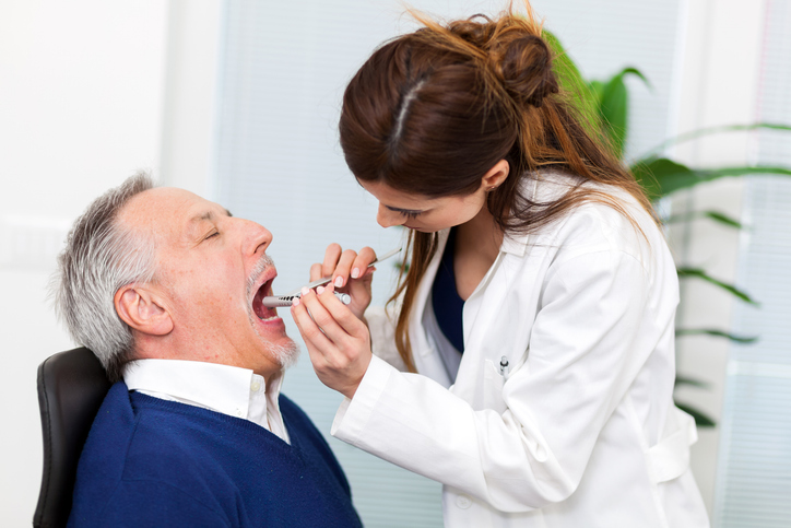 A dentist in a white coat examines an older man’s open mouth using a dental mirror and tool in a bright clinic, performing an oral cancer screening as she focuses on the patient’s teeth.