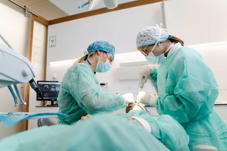 Two dental professionals wearing scrubs, masks, and caps are performing an oral surgery on a patient lying in a modern dental clinic. Dental equipment is visible around them.