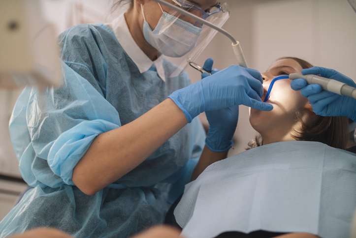 A dentist wearing protective gear and gloves uses dental instruments to examine a patient's teeth in a dental clinic, checking for cavities or signs that may require a root canal. The patient is lying back in the dental chair with a bib on.