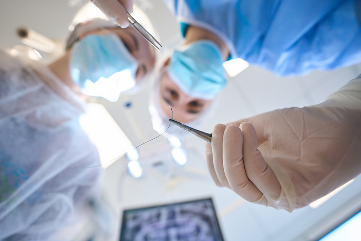 Two surgeons in masks and gloves, viewed from below, hold surgical instruments and a suture thread, preparing for suturing in a brightly lit operating room.