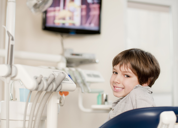 A young boy with brown hair smiles while sitting in a dentist’s chair. Dental equipment surrounds him, and TV viewing helps keep him relaxed as a television is mounted on the wall in the background.