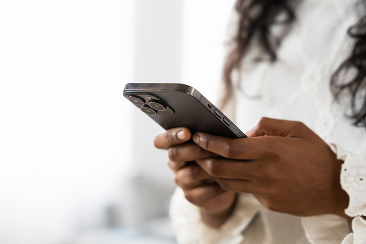 A person with dark skin and long hair holds a smartphone with both hands, likely text messaging, while wearing a white, long-sleeved, textured top. The background is softly blurred.