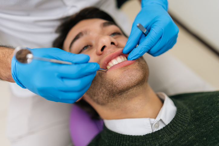 A person reclines in a dental chair while a dentist wearing blue gloves examines their teeth with dental instruments, possibly preparing for gum therapy.