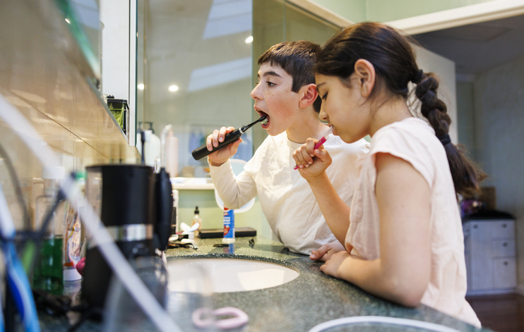 A boy and a girl stand at a bathroom sink brushing their teeth, practicing good oral hygiene; the boy uses an electric toothbrush while the girl uses a manual one. Both are focused on their task, with bathroom items visible on the counter.