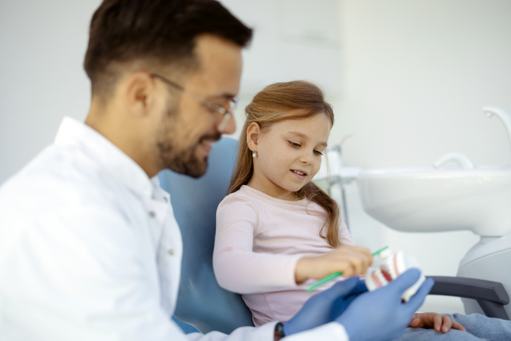 A male dentist demonstrates preventative dentistry to a young girl, showing her how to brush teeth using a dental model. Seated in the dental chair, she smiles while learning proper brushing techniques with a toothbrush.