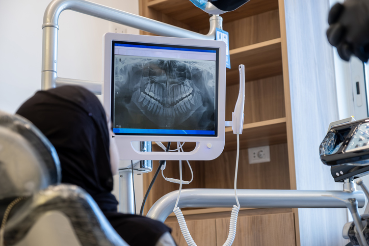 A dental X-ray image used for radiographic diagnosis is displayed on a monitor in a dental office, with a patient sitting in the chair and dental equipment visible around them.