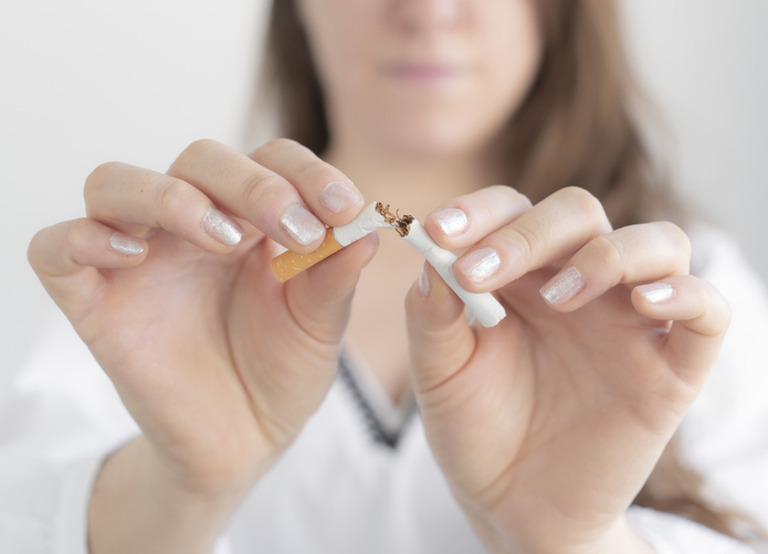 A woman breaks a cigarette in half with both hands, symbolizing smoking cessation. Her face is out of focus, while her hands and the broken cigarette are clearly visible in the foreground.