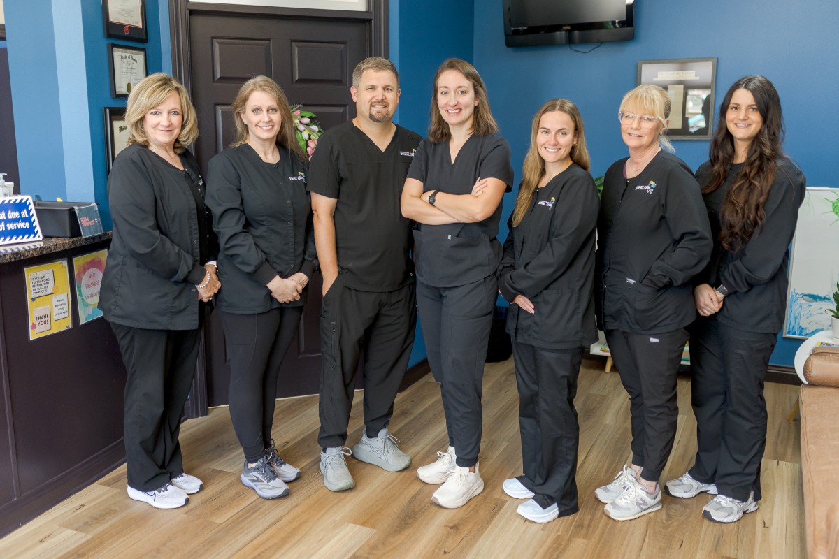 The Des Moines River Dental staff, dressed in black scrubs, stand smiling in a dental office with blue walls, wood flooring, and framed certificates displayed on the wall.