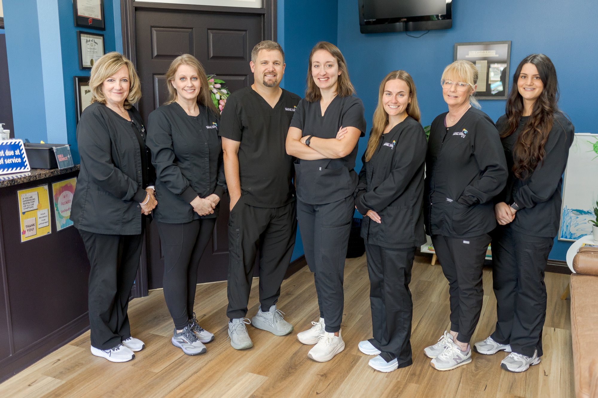Seven people dressed in black medical scrubs stand together and smile in the bright Des Moines River Dental office with blue walls and wooden flooring. Some have their arms at their sides, while one woman stands with arms crossed.