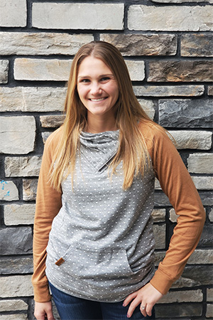 Hannah on our team, with long straight hair, smiles in front of a stone wall, wearing a gray polka-dot sweatshirt with tan sleeves and dark jeans.