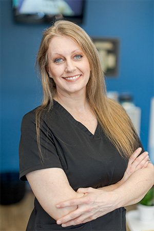 Brooke, a woman with long blonde hair wearing black scrubs, stands with her arms crossed, smiling confidently at the camera in a professional setting against a blue background.