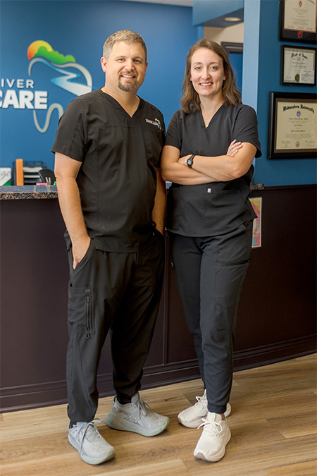Dr. Tyler and Dr. Emma Villiard, dressed in black scrubs and sneakers, stand smiling inside a dental office. Dr. Tyler stands with his hands in his pockets; Dr. Emma stands with arms crossed. Dental certificates and a logo are visible behind them.
