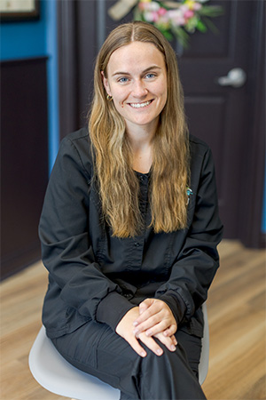 Taylor, a woman with long, light brown hair wearing a black outfit, sits on a chair with her hands folded in her lap, smiling at the camera. She is indoors with dark walls and wooden flooring in the background.